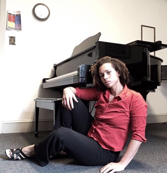 Jones half seated reclining in front of a piano. her right hand rests atop her knee